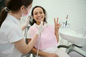 Deleitado misturado raça mulher paciente mostra uma Paz placa sentado dentro uma Dentistas cadeira e sorrisos com lindo cheio de dentes sorrir olhando às Câmera ao lado uma dental praticante realizando dental tratamento foto