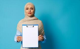 lindo árabe mulher vestindo uma bege hijab segurando uma prancheta com branco em branco Folha do papel foto