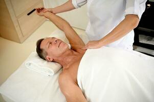 maduro caucasiano homem recebendo profissional terapêutico reabilitador massagem às bem estar clínica. corpo Cuidado, fisioterapia, esporte prejuízo reabilitação conceito, holístico Cuidado conceito foto