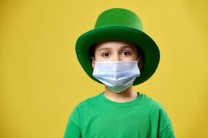 Garoto dentro verde irlandês duende chapéu e protetora médico mascarar poses para Câmera em uma amarelo fundo. santo patrick dia foto