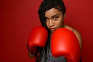 fechar-se retrato do concentrado jovem africano Esportes mulher boxer vestindo vermelho boxe luvas, fazer direto bater, isolado sobre vermelho fundo com cópia de espaço. jovem mulher durante cardio exercite-se foto