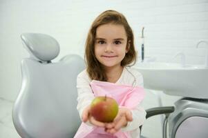 adorável pequeno menina sentado em uma dentista cadeira sorrisos olhando às Câmera e mostrando a maçã dentro dela estendido mãos depois de dental tratamento dentro moderno clínica. saudável comendo e oral higiene conceito foto