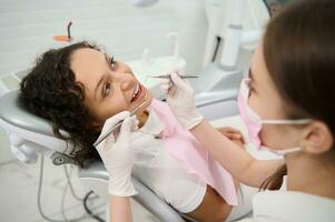 lindo mulher dentro Dentistas cadeira durante dental checar dentro odontologia clínica em borrado primeiro plano do dental higienista segurando inoxidável aço dental instrumentos e examinando pacientes oral cavidade foto