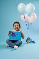 alegre menina criança fofa sorridente às a Câmera sentado em uma azul fundo Próximo para a alarme relógio e balões e segurando uma azul presente com uma Rosa arco foto