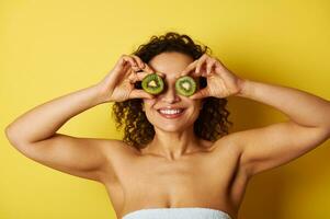 sorridente misturado raça muscular mulher cobertura dela olhos com kiwi metades, sorridente enquanto posando contra amarelo fundo foto