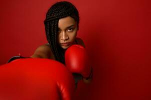 fechar-se retrato do concentrado jovem africano Esportes mulher boxer vestindo vermelho boxe luvas, fazer direto bater, isolado sobre vermelho fundo com cópia de espaço. Preto mulher boxer soco para Câmera foto