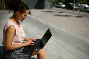 lado retrato do ocupado freelance mulher, iniciante , escritório trabalhador usando computador portátil sentado em passos durante café pausa. escritório trabalhador em almoço pausa dentro urbano fundo foto