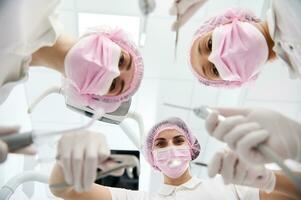 inferior Visão do fêmea Dentistas e dental assistente dentro Rosa protetora mascarar segurando estéril Ferramentas para uma dental exame e inclinado para uma paciente . foco em dental instrumentos foto