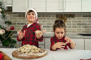 alegre Deleitado crianças olhando às Câmera e mostrando seus mãos dentro a massa. adorável crianças tendo diversão, jogando com massa enquanto Aprendendo culinária às casa durante Natal feriados foto