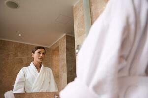 espelho reflexão do a atraente confiante meio envelhecido morena mulher vestindo roupão de banho olhando às ela mesma em pé dentro banheiro durante manhã higiene rotina foto