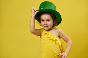 fofa pequeno menina com verde duende irlandês chapéu sorrisos para Câmera em pé em uma amarelo fundo. santo patrick's dia foto