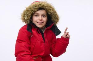 bonito alegre pré-adolescente, escola envelhecido caucasiano Garoto vestindo brilhante vermelho parka com capuz, sorridente cheio de dentes sorrir olhando às Câmera e apontando com dedo em uma cópia de espaço do branco fundo foto