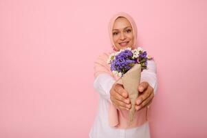 uma lindo simples ramalhete do Prado flores silvestres dentro roxa tons, embrulhado dentro Castanho construir papel dentro a mãos do a atraente sorridente muçulmano mulher vestindo uma hijab. isolado Rosa fundo cópia de espaço foto