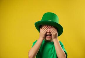 pequeno rindo Garoto dentro verde irlandês duende chapéu cobre dele olhos com dele Palmeiras. santo patrick dia em amarelo fundo com cópia de espaço foto