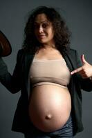alegre grávida mulher apontando dela dedo em dela grávida barriga, sorridente olhando às Câmera, isolado estúdio fundo foto
