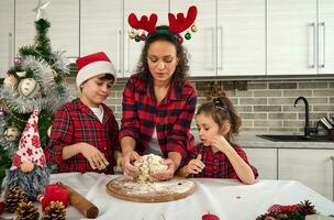 pequeno crianças ajudando seus mãe para amassar uma massa em uma de madeira quadro, cozinhando às casa cozinha para Natal tradicional eventos. lindo mãe ensino dela filho e filha quão para preparar a massa foto