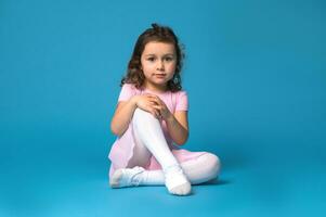 retrato do uma fofa pré escola menina bailarina vestindo Rosa vestir , sentado em azul fundo e posando para a Câmera foto