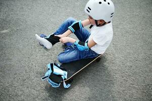 desportivo Garoto dentro segurança capacete sentado em skate em Parque infantil asfaltos e colocando em protetora joelho almofadas foto