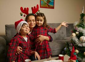 feliz mãe com chifre aro abraços dela adorável crianças vestido dentro vermelho e verde xadrez roupas e santa chapéu em dela filhos cabeça, a comemorar Natal festa às casa dentro família círculo foto