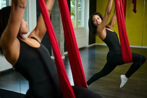 em forma bonita jovem mulher vestindo roupa de esporte fazendo mosca ioga alongamento exercícios dentro ginástica Treinamento dentro ioga estúdio com grande janelas. aéreo aero mosca ginástica em vermelho maca com anti-gravidade foto