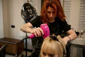 frente Visão do uma ruivo mulher profissional cabeleireiro com tatuado braços dentro uma rigoroso Preto uniforme pentear, endireitar e secagem a Loiras cabelo do beleza salão cliente usando uma Rosa secador de cabelo foto