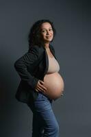 alegre jovem grávida mulher dentro azul jeans, cinzento blazer e bege sutiã, segurando dela barriga, sorridente olhando às Câmera foto