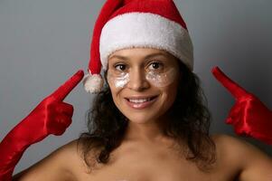 atraente morena dentro santa traje pontos dedos em olho remendos em dela face, sorrisos olhando às Câmera. publicidade para beleza salões para Natal e Novo ano Sorteios. spa, pele Cuidado conceito foto