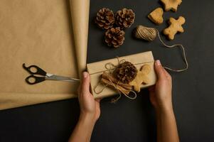 mãos segurando feito à mão Natal presente caixa embrulhado dentro Castanho construir papel decorado com pinho cones e canela deitado em uma Preto superfície com uma Pão de gengibre homem e biscoitos, corda e tesoura. plano deitar foto