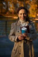 retrato do uma alegre africano mulher com Leve embora copo do café e uma mão livro sorridente olhando às Câmera em a outono parque lago fundo com queda folhas. outonal ao ar livre lazer. lazer foto