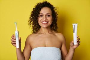 lindo mulher embrulhado dentro uma branco banho toalha segurando uma pasta de dentes e a elétrico escova de dente e sorridente às a Câmera com cheio de dentes sorrir foto