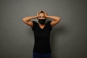 isolado retrato em cinzento fundo do a espantado atônito mulher dentro Preto protetora médico mascarar colocando dela mãos em dela cabeça olhando às Câmera. Preto Sexta-feira conceito foto