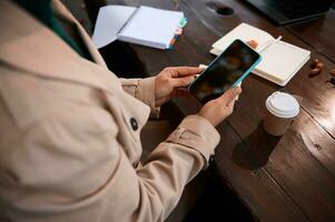 irreconhecível pessoa mãos usando uma inteligente telefone com em branco Preto tela para cópia de espaço em a fundo do uma de madeira mesa com aberto bloco de anotações e caderno e delicioso café da manhã foto