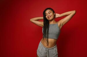 retrato do uma lindo alegre africano mulher com à moda dreadlocks, sorrisos olhando a parte, de lado posando contra vermelho fundo com braços atrás dela cabeça. cópia de espaço foto