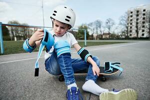 adorável Garoto skatista dentro segurança capacete e engrenagem sentado em de madeira skate em Parque infantil asfalto foto