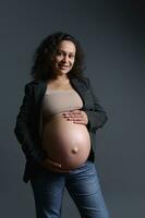 retrato do uma grávida mulher dentro cinzento blazer e bege sutiã, acariciando dela barriga, sorridente e olhando com confiança às Câmera foto