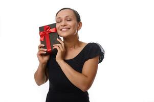 atraente mulher com fechadas olhos vestido dentro Preto poses para Câmera contra branco fundo, com Preto presente caixa perto dela face, sorrisos cheio de dentes sorriso, desfrutando presente. Preto sexta-feira, aniversário conceito foto