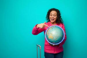 alegre delicioso mulher dentro Rosa Jaqueta pontos com dedo para viagem destino em a mapa do uma globo dentro dela mãos. conceito do turismo, viagem por aí a mundo com cópia de de Anúncios espaço em azul fundo foto