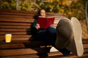 a Principal foco em a único do Esportes tênis do uma borrado mulher em repouso em uma de madeira Banco dentro a parque e lendo uma livro e bebendo café. desfrutando uma final de semana ao ar livre dentro outono natureza fundo foto