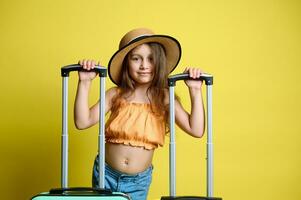 caucasiano feliz criança, pequeno menina dentro oculos de sol, Palha chapéu e laranja principal, posando com malas, isolado em amarelo foto