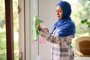 retrato do uma confiante linda jovem árabe muçulmano mulher com coberto cabeça dentro azul hijab lavando janelas, removendo manchas e limpando com trapo enquanto desfrutando Primavera limpeza dentro casa. cópia de espaço para de Anúncios foto