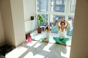 mulher e garota, mãe e dela filha praticando ioga em uma ginástica esteira às casa em uma lindo ensolarado dia. desfrutando união e meditação foto