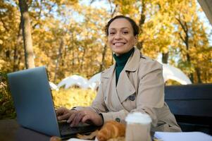 atraente mulher com lindo sorriso, o negócio pessoa, trabalhador autonomo desfrutando ao ar livre remotamente trabalhar, trabalhando em computador portátil dentro a carvalho Arvoredo com construir papel copo do café e cozido croissant em primeiro plano foto