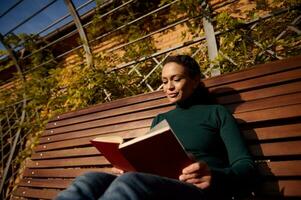 relaxado confiante mulher dentro casual jeans e verde estacionar lendo uma livro sentado em uma de madeira Banco desfrutando uma caloroso ensolarado outono final de semana dentro país parque, longe a partir de a encontrão e urgência do a cidade foto