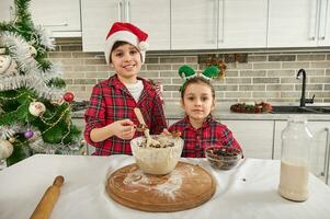 lindo caucasiano crianças, bonito alegre Garoto e dele mais jovem irmã, fofa bebê menina olhando às Câmera em pé de uma cozinha mesa e cozinhando Natal bolos massa foto