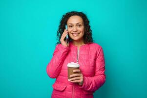 encantador impressionante mulher dentro brilhante Rosa Jaqueta detém descartável cartão copo com quente café e sorrisos cheio de dentes sorriso, falando em célula telefone e olhando às Câmera, isolado em azul fundo cópia de espaço foto