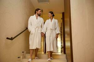 lindo amoroso recém-casado casal, vestindo branco roupões de banho, desfrutando seus lua de mel juntos dentro luxo spa recorrer, segurando mãos Como elas andar baixa a escadas do a bem estar spa complexo foto