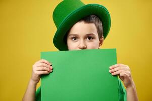 bonito Garoto vestido dentro uma duende com uma verde irlandês boné cobre dele face com uma Folha do verde papel com cópia de espaço. santo patrick's dia conceito. foto