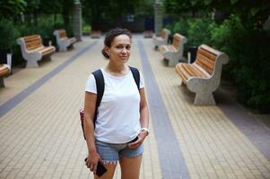 lindo mulher dentro branco brincar camiseta e azul jeans, sorridente olhando às Câmera, em pé dentro a cidade parque quadrado foto