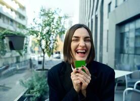 alegre espantado mulher segurando crédito cartão com verde chromakey perto dela face, sorrisos cheio de dentes sorrir posando contra a fundo do moderno arranha-céus edifícios fundo. o negócio e finança conceito foto