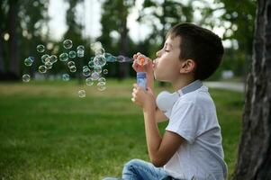 adorável bonito pré-adolescente criança Garoto sopro iniciando Sabonete bolhas em a parque, em repouso durante dele escola lazer foto